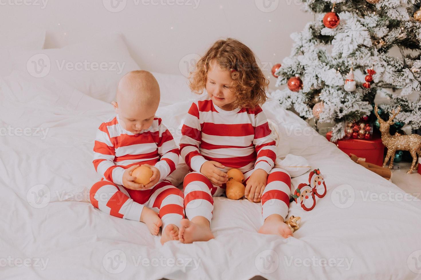 children in red and white pajamas try on funny glasses with Santa Claus sitting in bed. lifestyle. brother and sister celebrating Christmas. boy and girl are playing at home. High quality photo