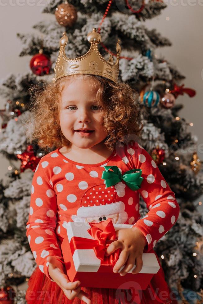 little curly-haired girl in a carnival dress hid her face behind shiny Christmas tree toys stars. child in a red dress with a Santa print on the background of a Christmas tree. High quality photo