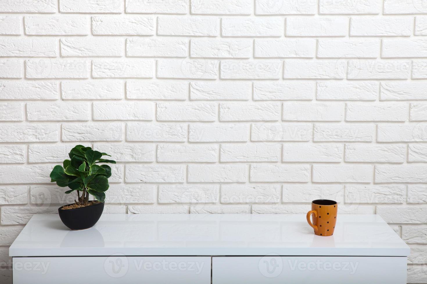 Cup and green pot on a white table near the white wall. Empty workplace in the room. White background mock up. photo