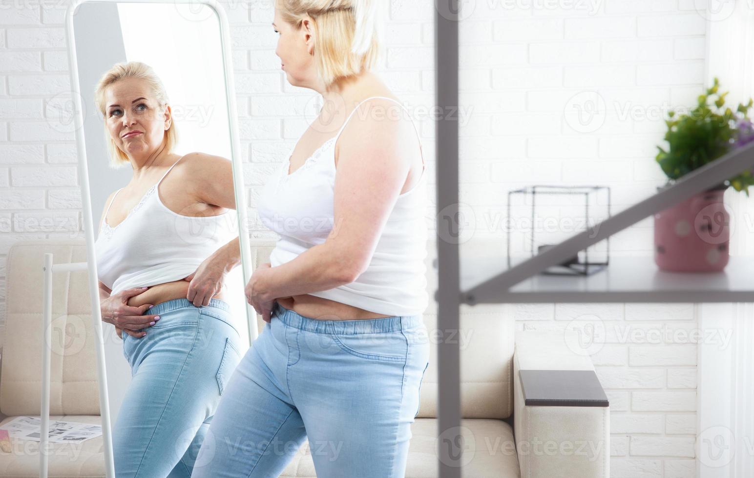 Displeased middle aged woman looking at herself in the mirror and pinching stomach fat in domestic interior. Sad plus size woman looking her reflection in the mirror with sorrow at home. photo