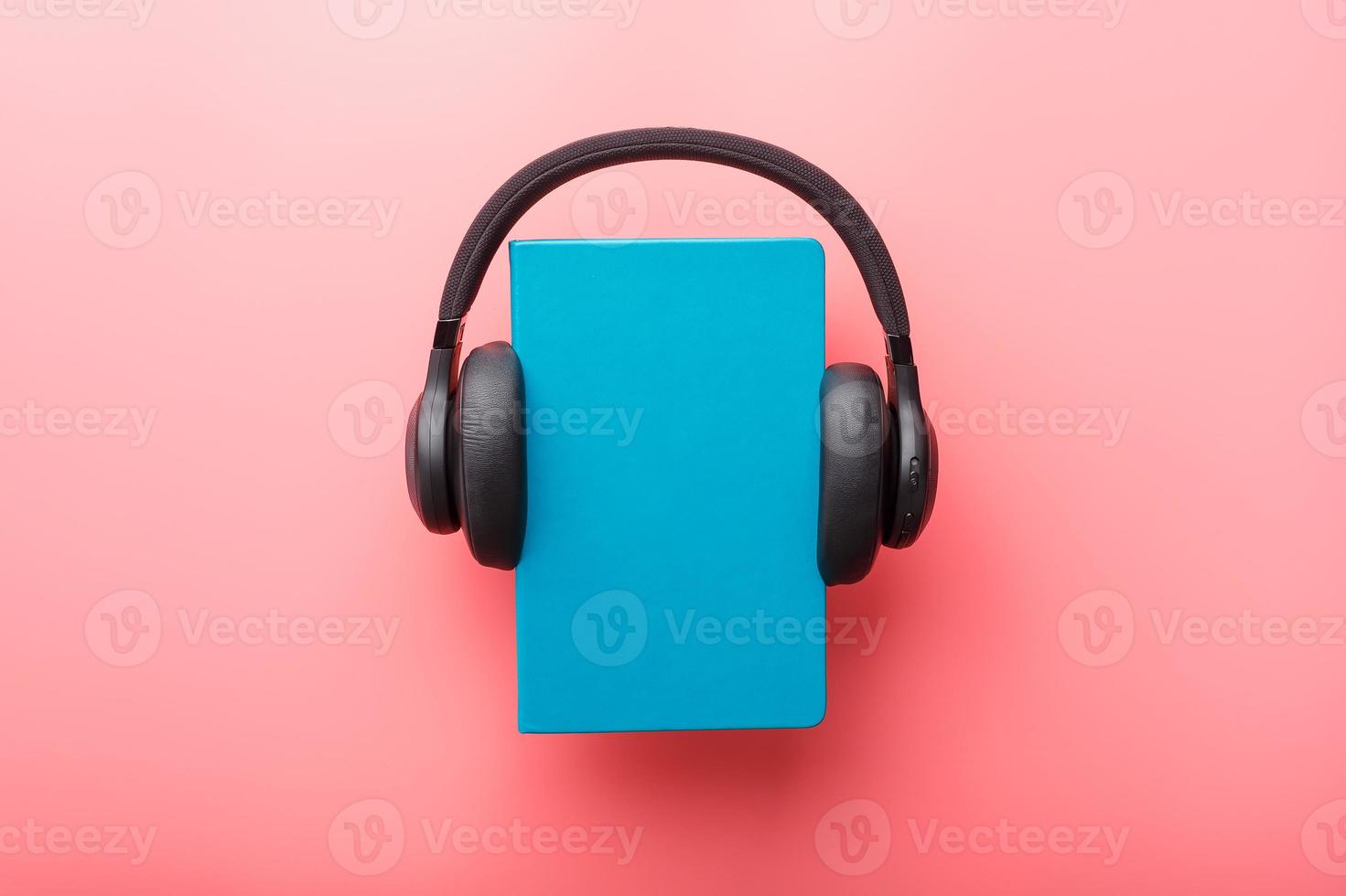 Headphones are worn on a book in a blue hardcover on a pink background, top view. photo