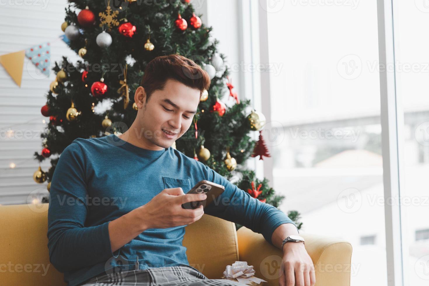 A single man smiling while looking his phone in his living room decorated with Christmas trees photo