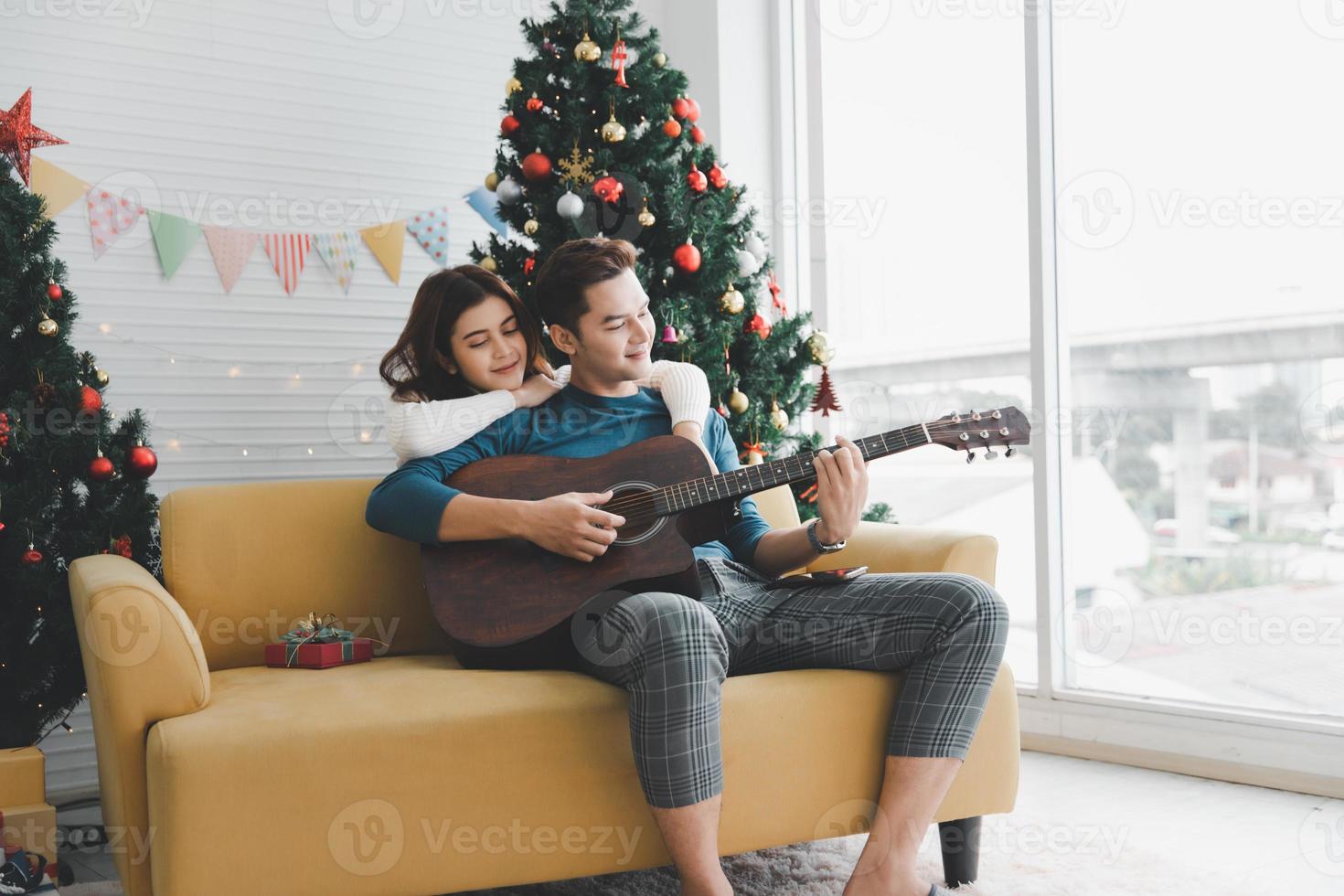 un hombre tocando la guitarra a una mujer amante durante la celebración de las vacaciones de navidad juntos en la sala de estar con decoración de adornos de árboles de navidad foto