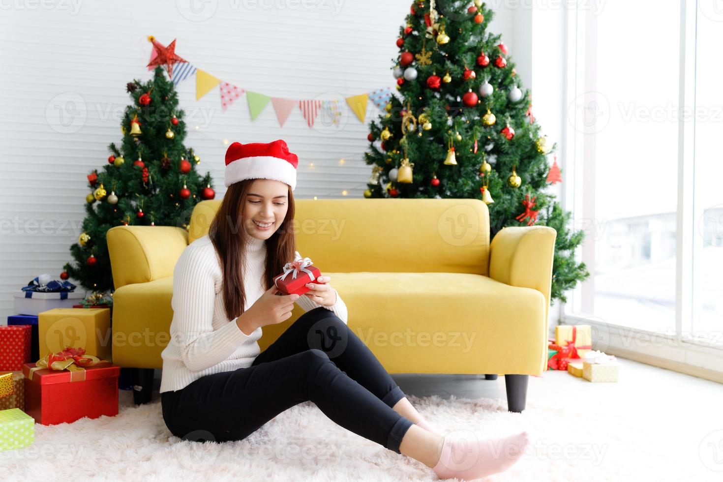 Smiling brunette woman in sweater and Santa hat sitting on the floor in living room holding small gift box and rejoices over Christmas tree decorative background photo