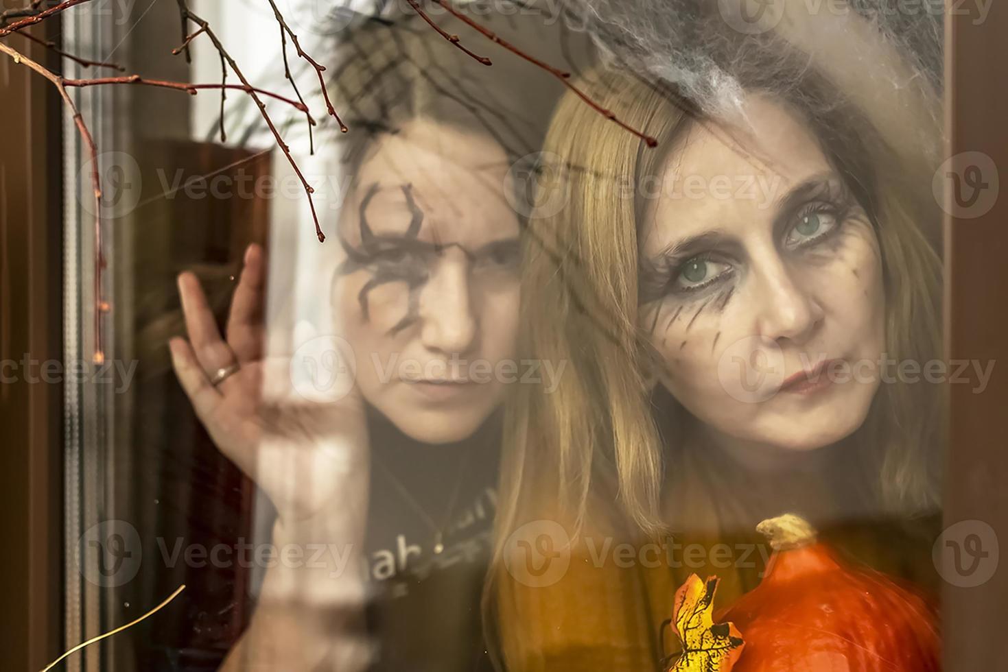 Two female witches look through a spider web ominously out of the window against the backdrop of Halloween decorations. Masquerade, Halloween party photo