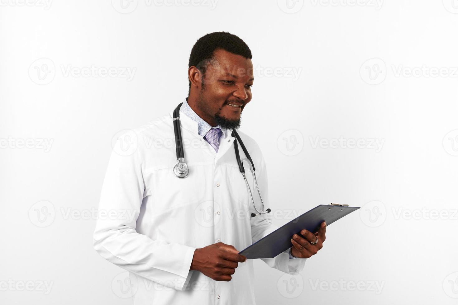 Smiling black bearded doctor man in white robe with stethoscope holds medical chart on clipboard photo