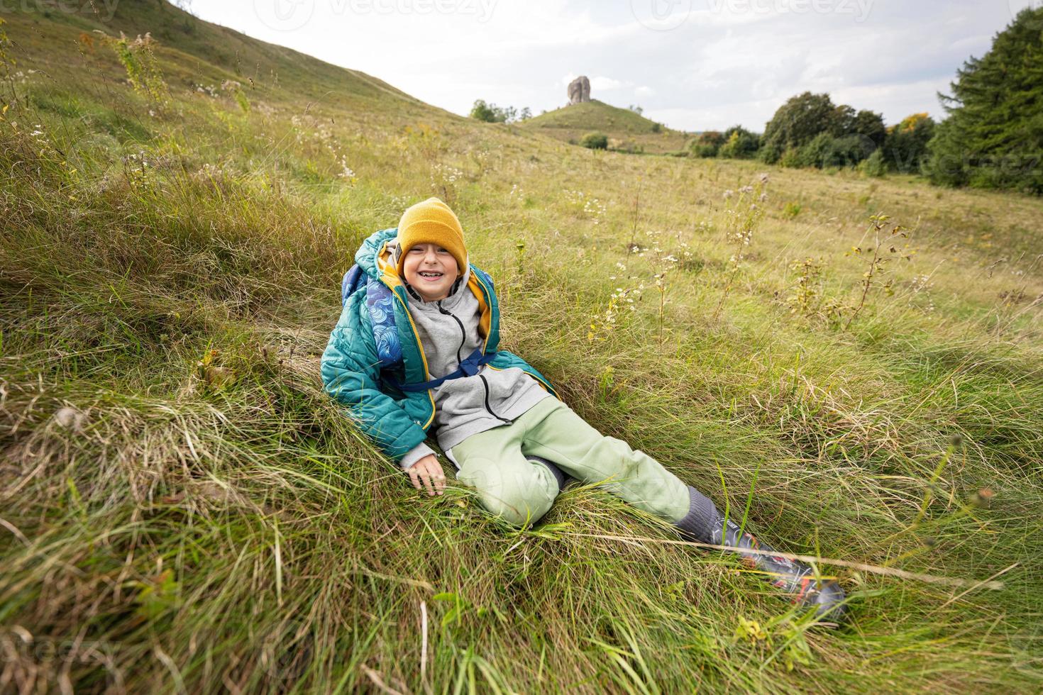 Smiling boy sitting at autumn hill wearing a green jacket, yellow hat, with backpack and rubber boots. photo