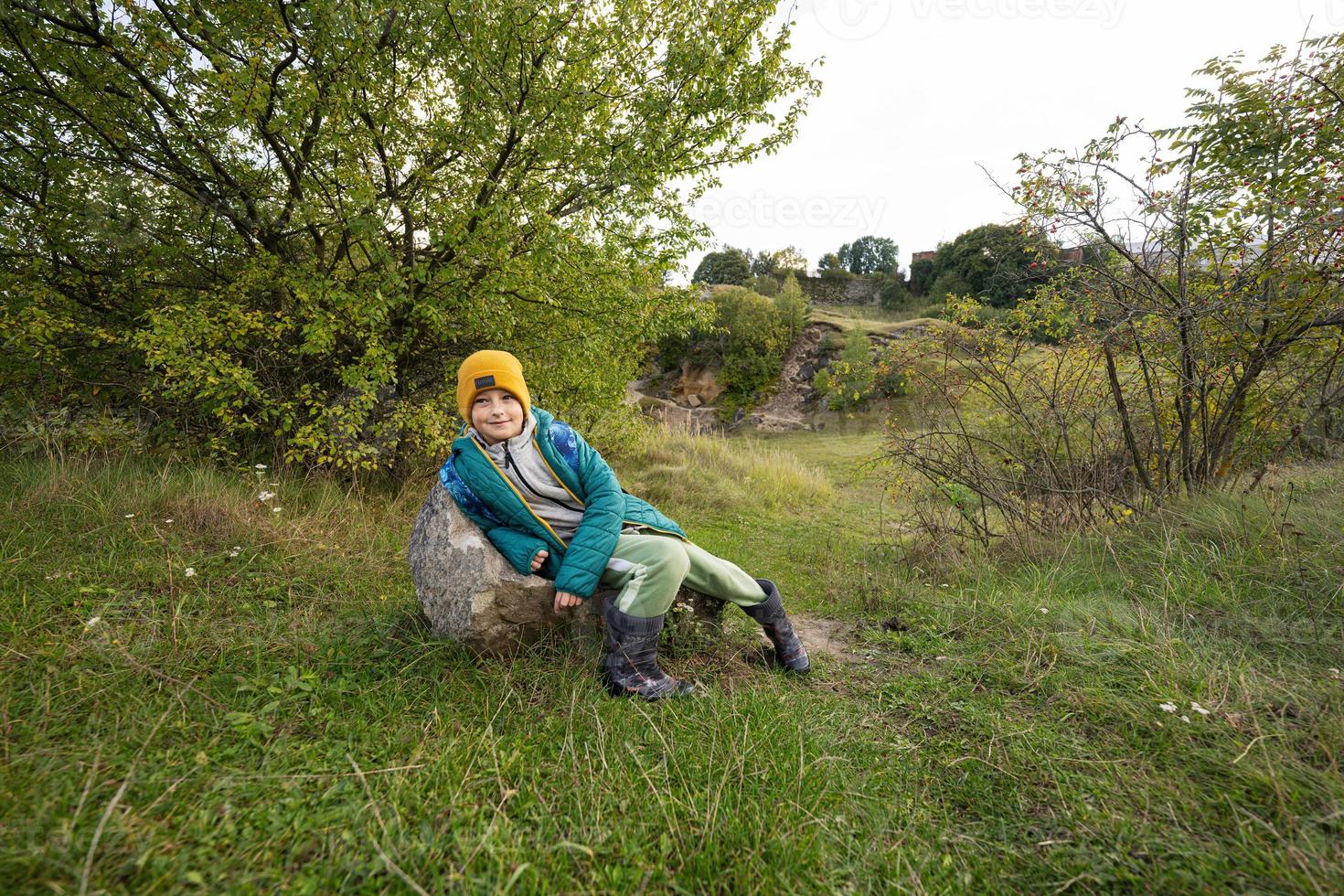 Cute boy sit on stone at autumn hill wearing a green jacket, yellow hat, with backpack and rubber boots. photo