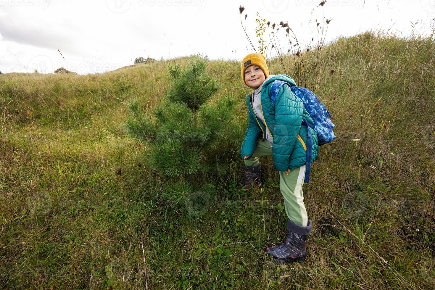 Cute boy stand near pine tree at autumn hill wearing a green jacket, yellow hat, with backpack and rubber boots. photo