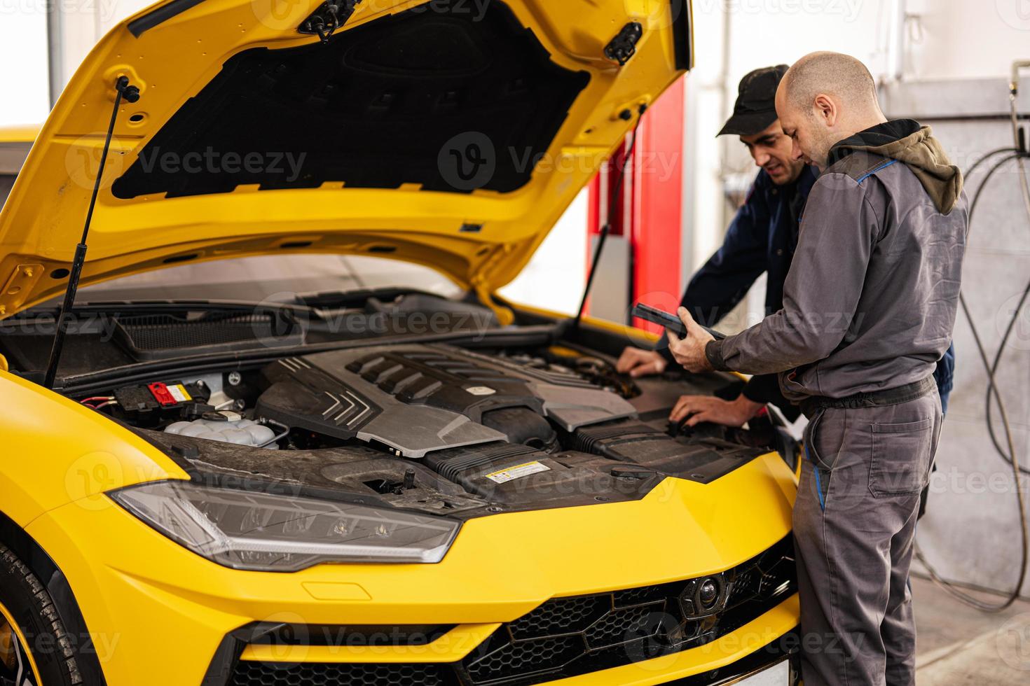 Two mechanics stand with tbalet against open hood of yellow sport car suv. photo