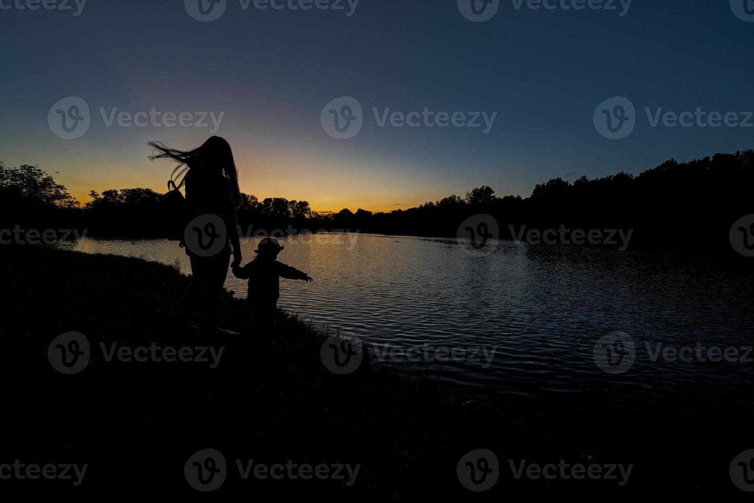 Silhouettes of mother and daughter holding hand against lake in sunrise dawn. photo