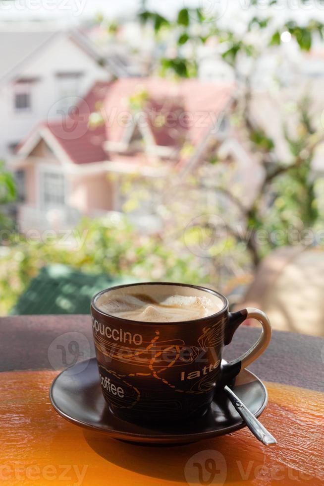 Cup with hot milk coffee on a colorful wooden table in a cafe on background of city view. Dalat, Vietnam. photo