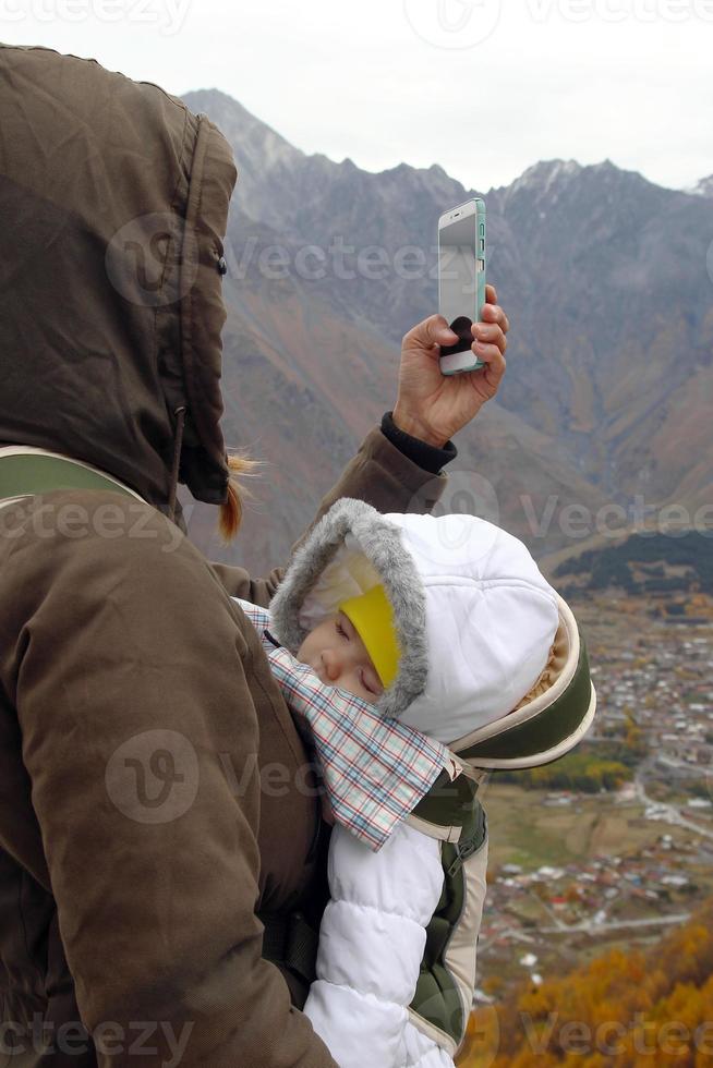 Portrait of young mother with her little sleeping girl in ergo carrier on a background of autumn mountains. Woman is taking a photo from smartphone.