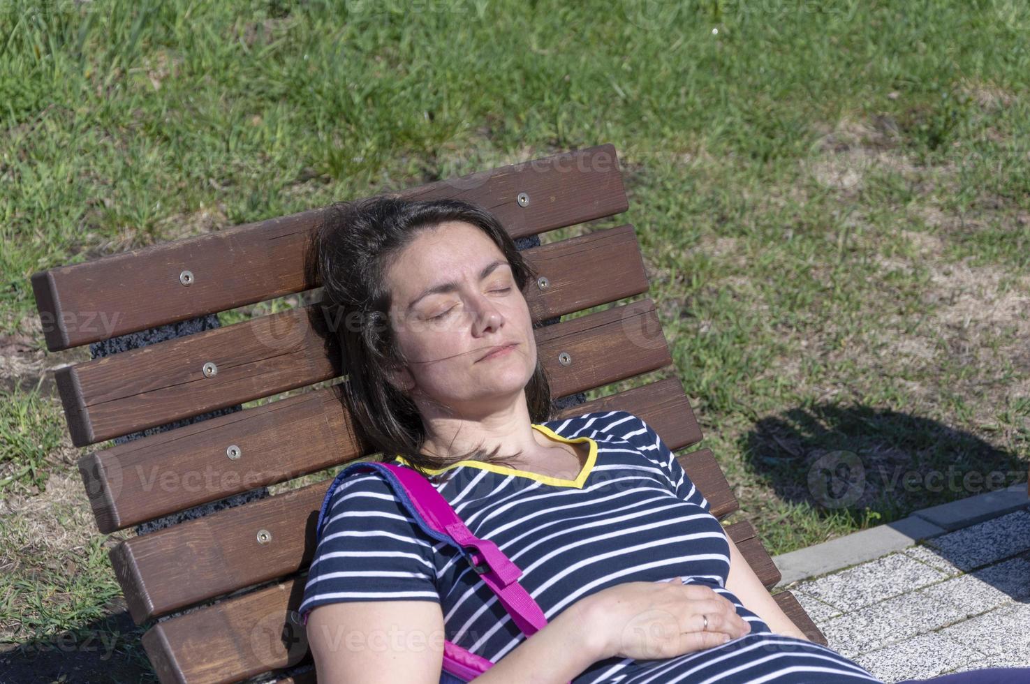 a young brunette woman lies on a wooden chaise longue in a city park on a sunny day photo