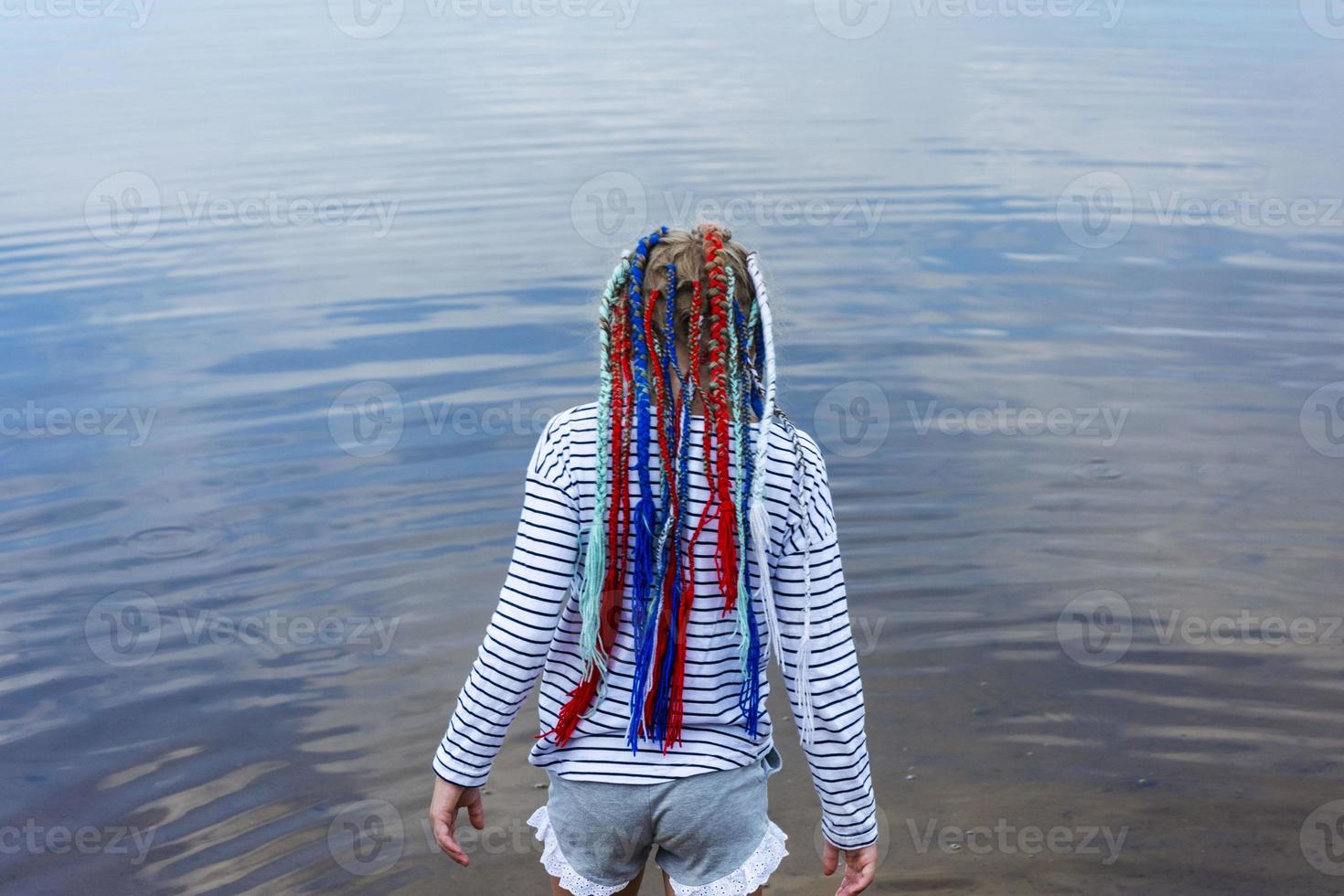 A teenage girl with bright multicolored ribbons braided in pigtails, a modern youth hairstyle, a girl goes into the water of the lake photo