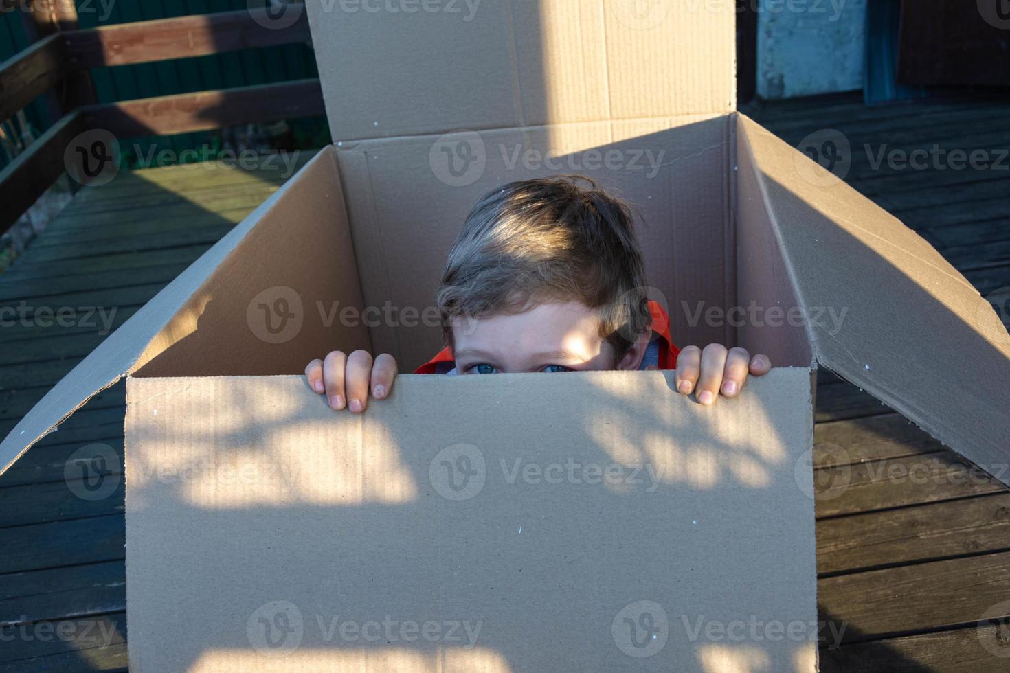 lindo niño europeo asomándose de una caja de cartón, regalo, sorpresa, vida sin hogar, empaque, juego de escondite foto