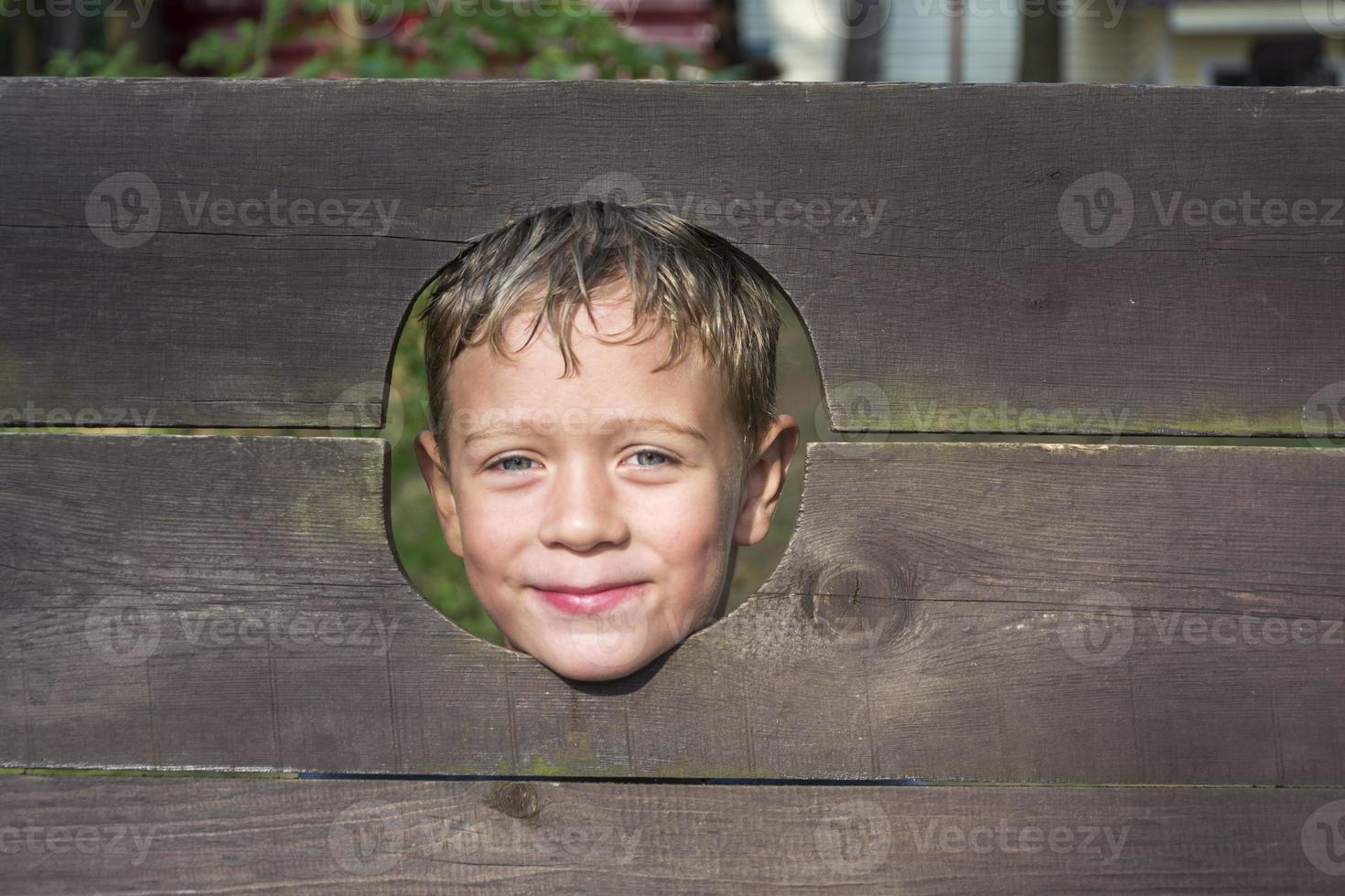 A cute Caucasian boy looks through a hole in a wooden fence and smiles photo