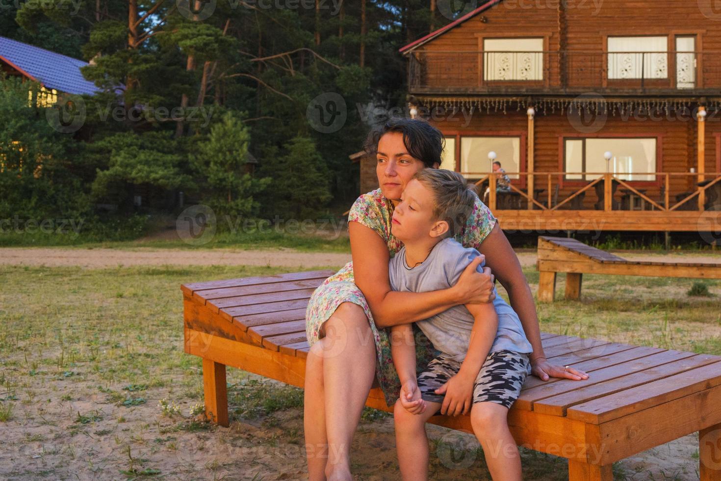 mom hugs her son sitting on a wooden bench on vacation on the evening beach in the light of the setting sun photo