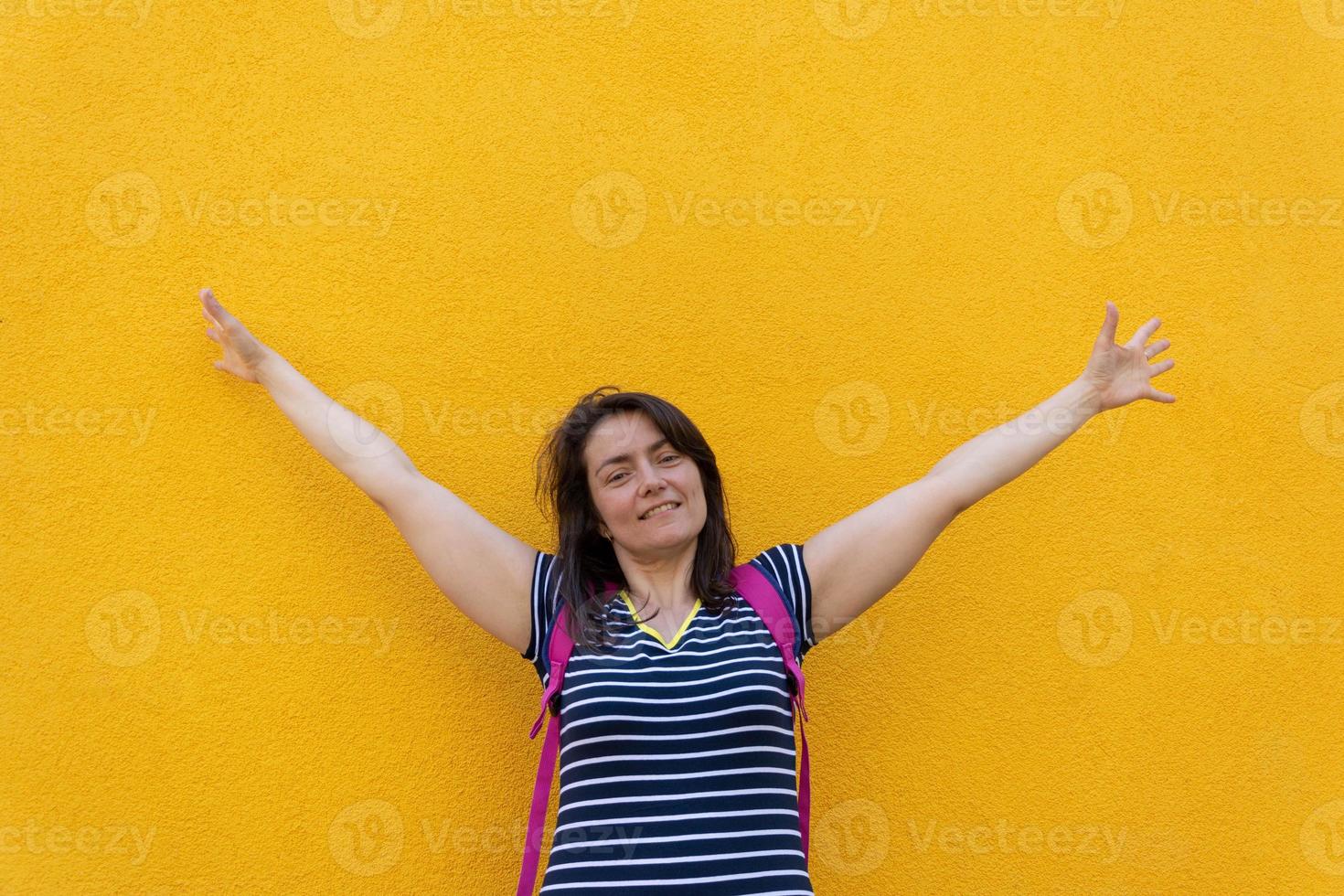 Portrait of a smiling brunette woman with a backpack, standing on a yellow background, spread her arms photo