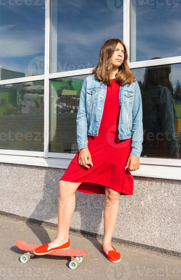 linda chica con un vestido rojo y una chaqueta de mezclilla con una patineta cerca de una ventana de una tienda de vidrio, zapatos deportivos rojos foto