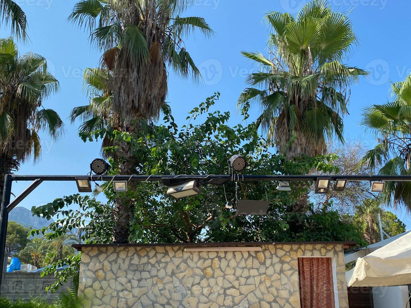 Audio visual scaffolding with concert spotlights and speakers, at an outdoor music festival, with a palm tree and blue sky in the background, and space for text on left photo