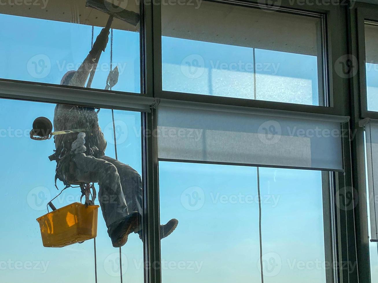 un trabajador de lavado de ventanas masculino, escalador industrial cuelga de un edificio alto, rascacielos y lava grandes ventanas de vidrio para limpieza en lo alto de una gran ciudad foto