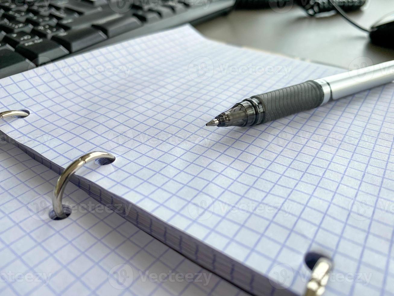 A writing pen rests on a notepad with squared paper sheets on a work desk with stationery in a business office photo