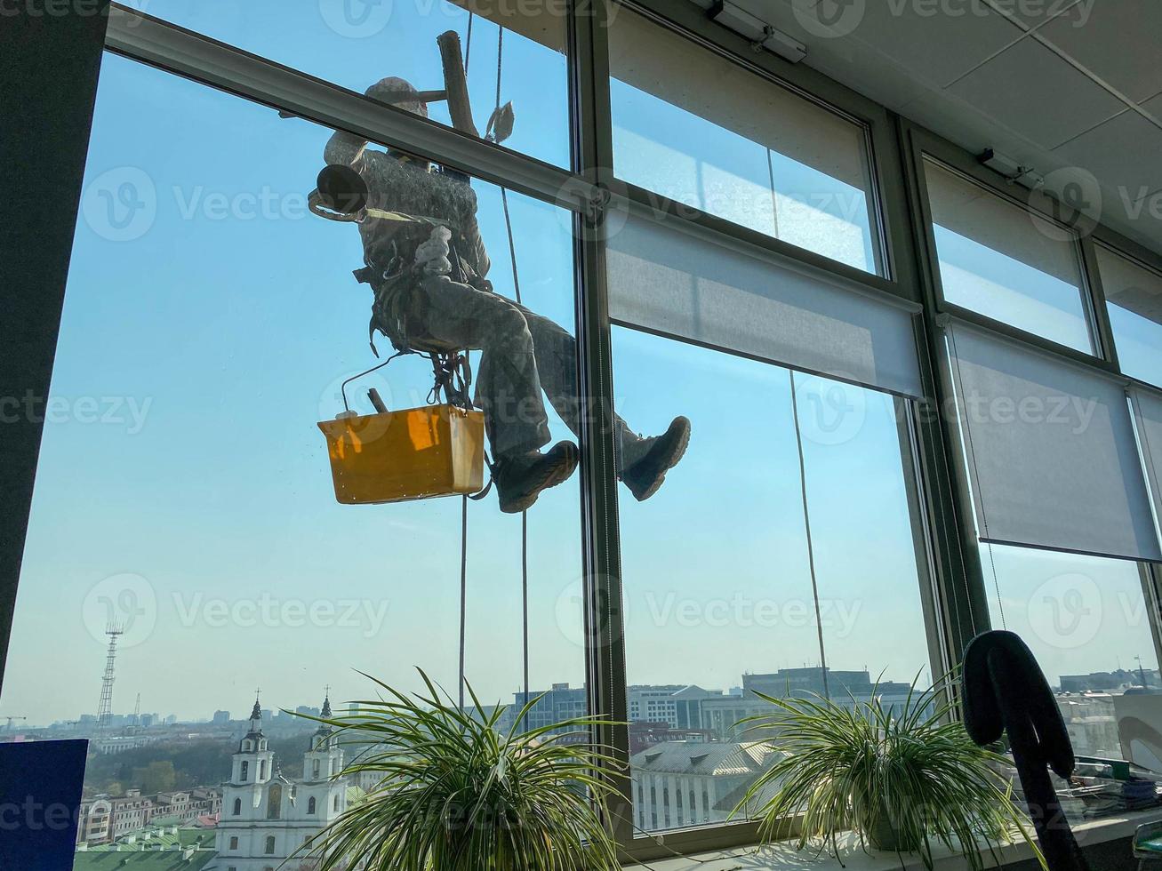 A male window washer worker, industrial climber hangs on a tall building, skyscraper and washes large glass windows for cleanliness high above a large city in an office business building photo