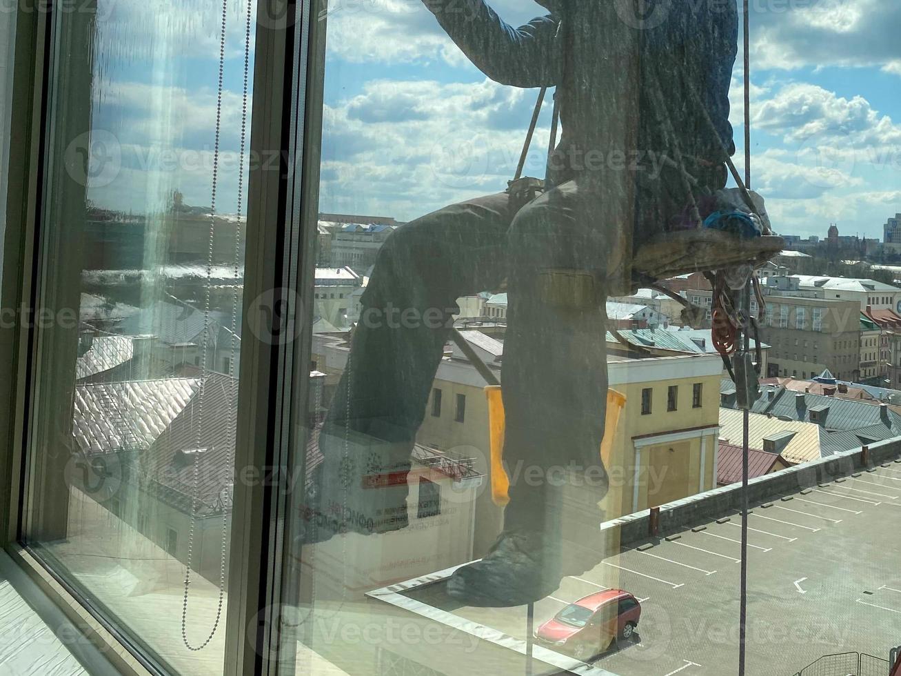 un trabajador de lavado de ventanas masculino, escalador industrial cuelga de un edificio alto, rascacielos y lava grandes ventanas de vidrio para limpieza en lo alto de una gran ciudad foto