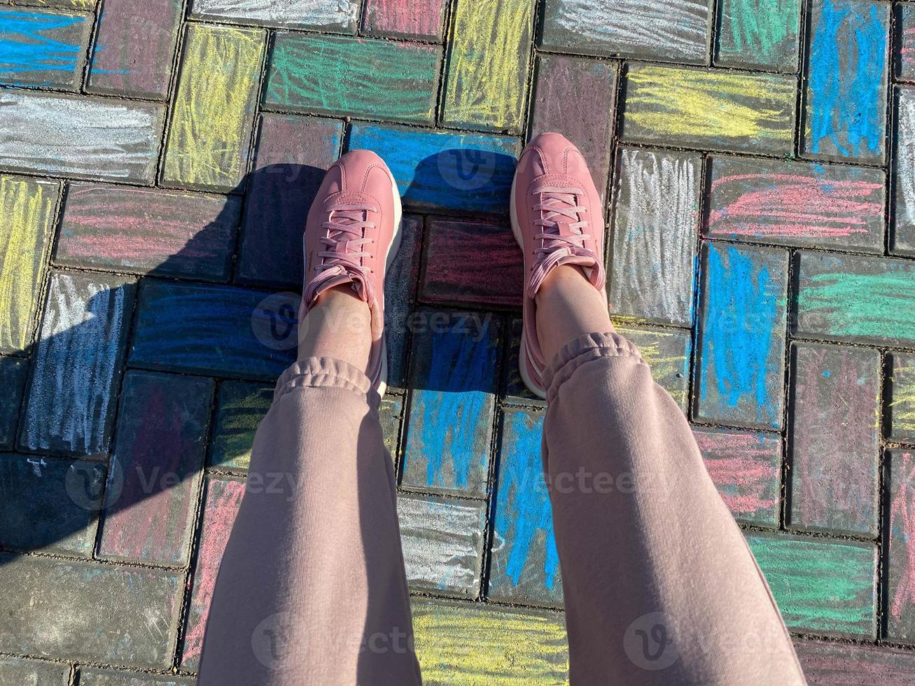 Beautiful female legs in pink sneakers and fashionable pants on a background of concrete paving slabs painted with joyful children's colorful bright crayons photo