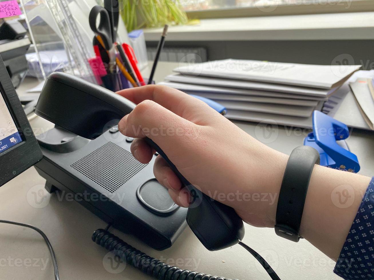 A man's hand in a shirt holds a landline phone receiver on a work desk with office supplies in a business office photo