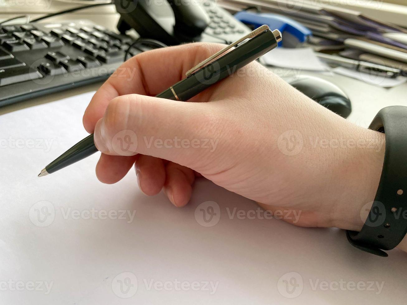A man's hand in a shirt and with a fitness bracelet holds a pen and writes on the table at the office table with a computer with a keyboard. Business work photo