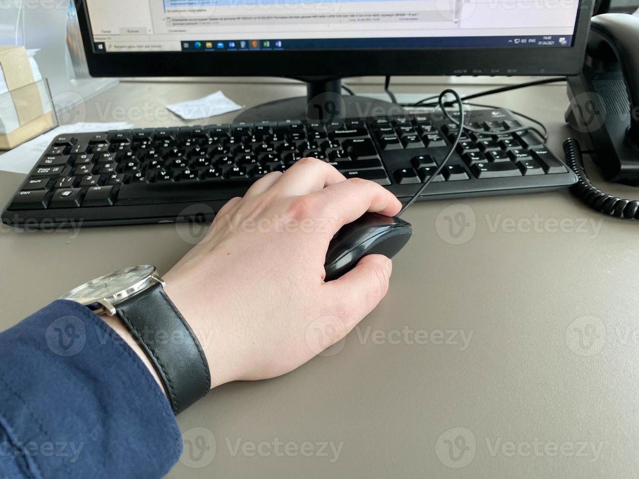 A man's hand with a shirt and a clock holds a computer mouse on the table at the office table with a computer with a keyboard. Business work photo
