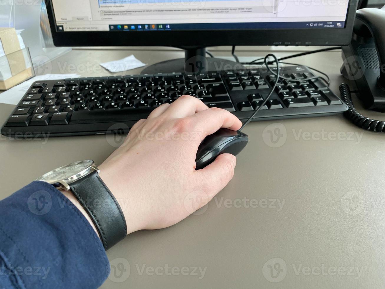 la mano de un hombre con una camisa y un reloj sostiene un ratón de computadora en la mesa de la oficina con una computadora con teclado. trabajo de negocios foto