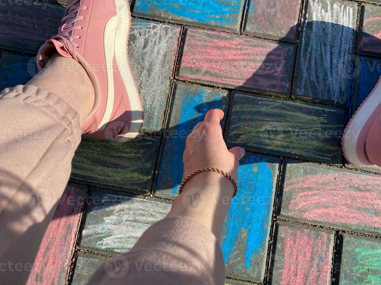 Beautiful female legs and hands in pink sneakers and fashionable pants on a background of paving slabs painted with children's colorful bright crayons photo