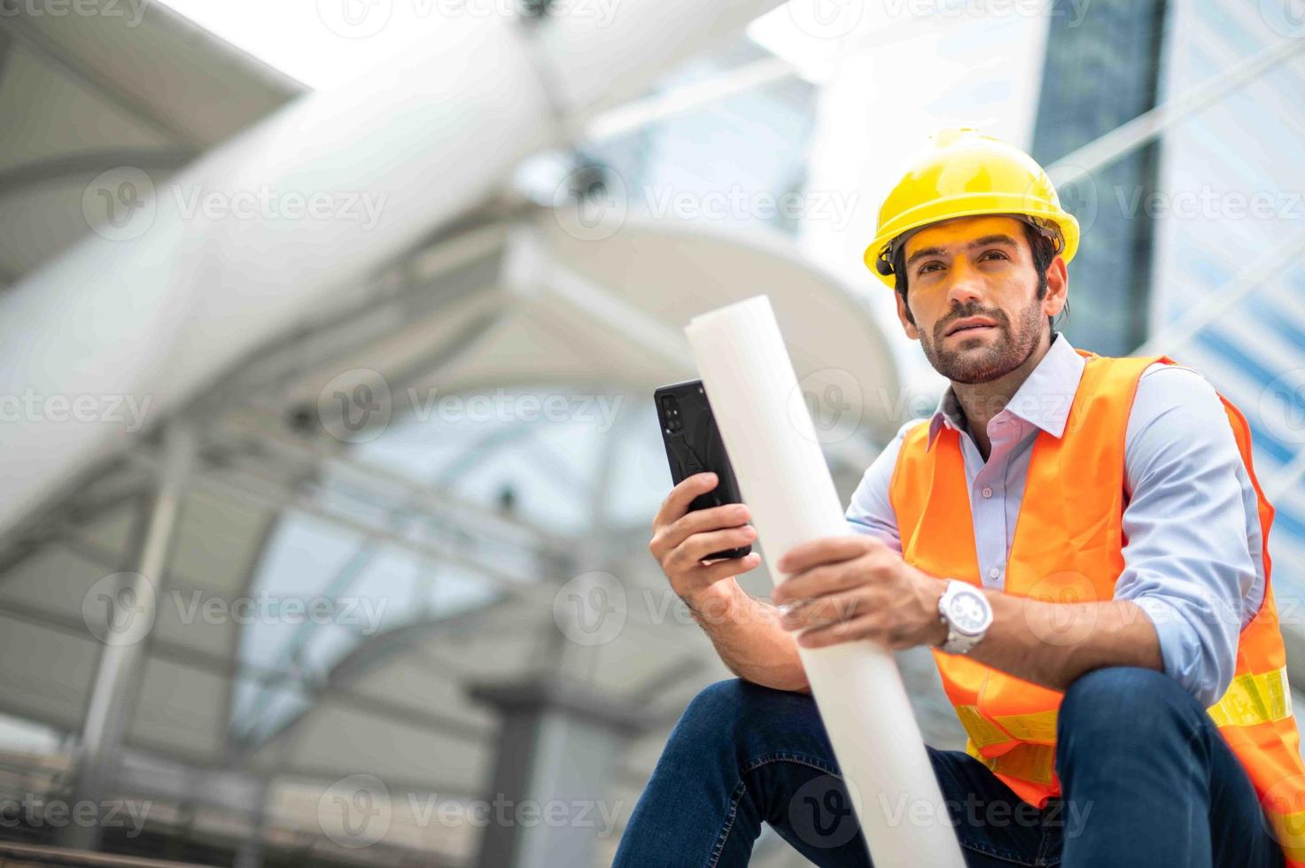 Caucasian man engineer use a smartphone for talking, wearing orange vest and big hard hat, and the other hand holding the white floor plan in the site work of the center city. photo