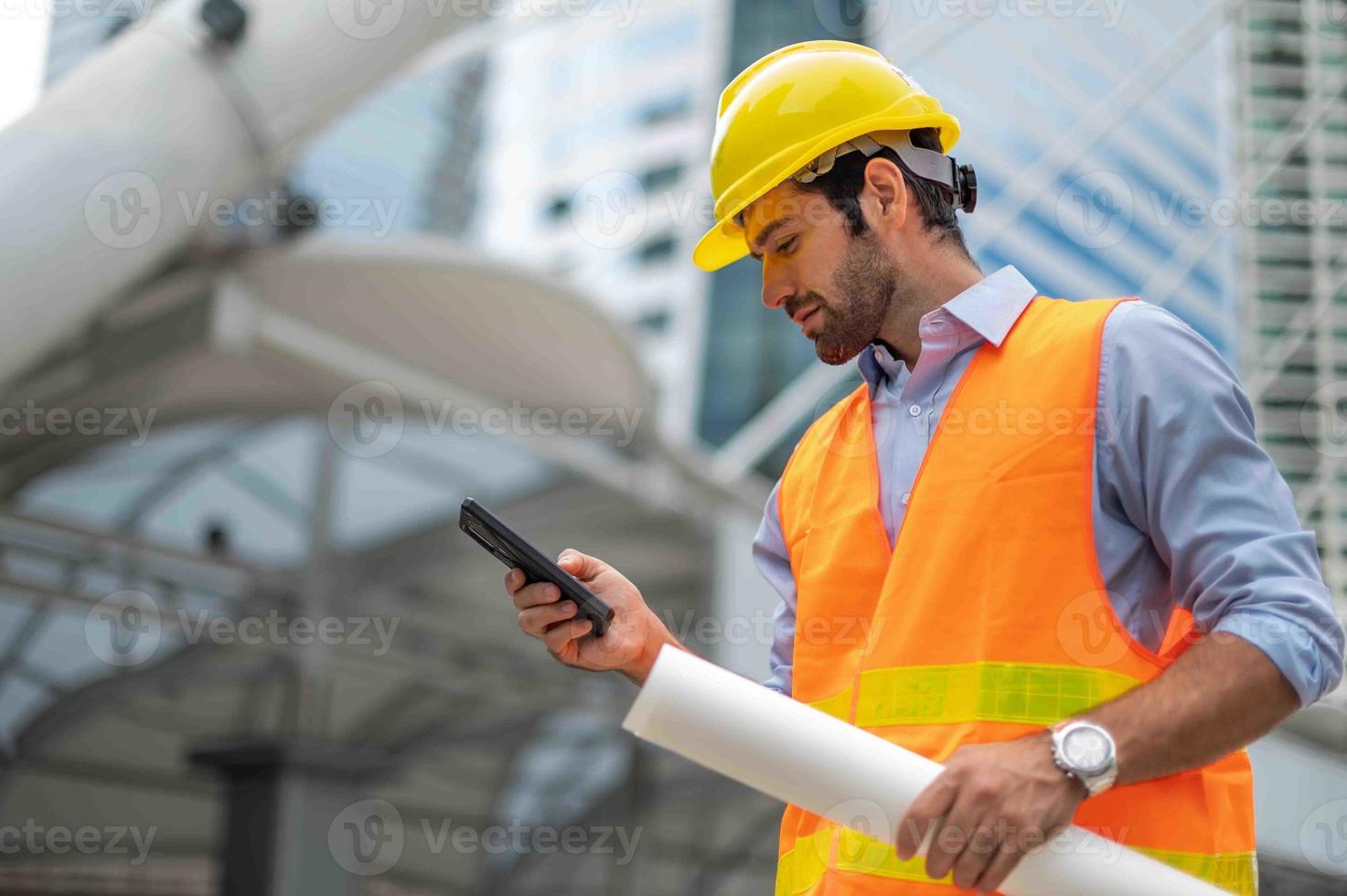 el ingeniero caucásico usa un teléfono inteligente para hablar, usa chaleco naranja y un gran sombrero duro, y la otra mano sostiene el plano de planta blanco en el sitio de trabajo del centro de la ciudad. foto