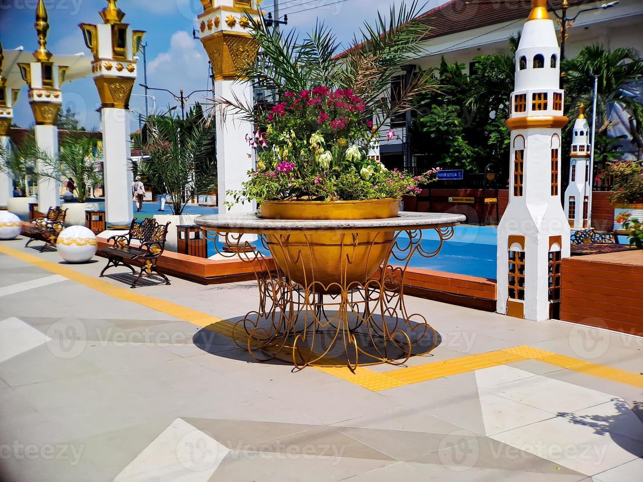 Large flower pot with golden iron carvings, Arabic style architecture, colorful flowers growing, bright blue clouds. photo