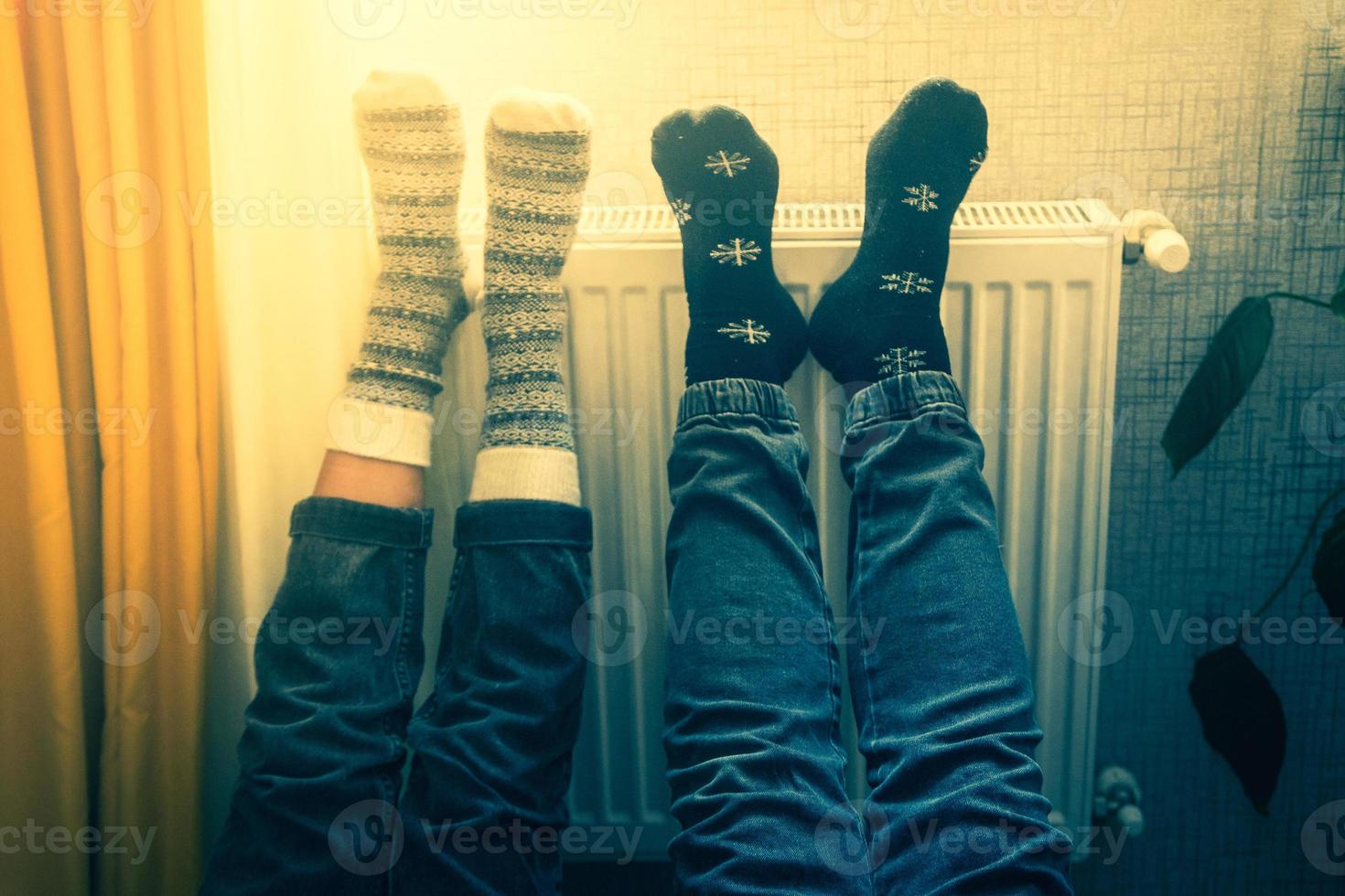 Couple hold legs up heating feet in cold home indoors on radiator in winter with cozy winter stylish woolen socks on. Valentines funny together warm feet by radiator by window photo