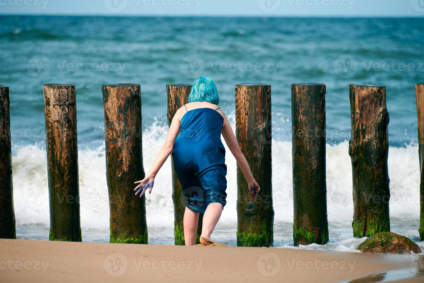 Blue-haired woman performance artist in blue dress standing on beach holding paint brush, back view photo