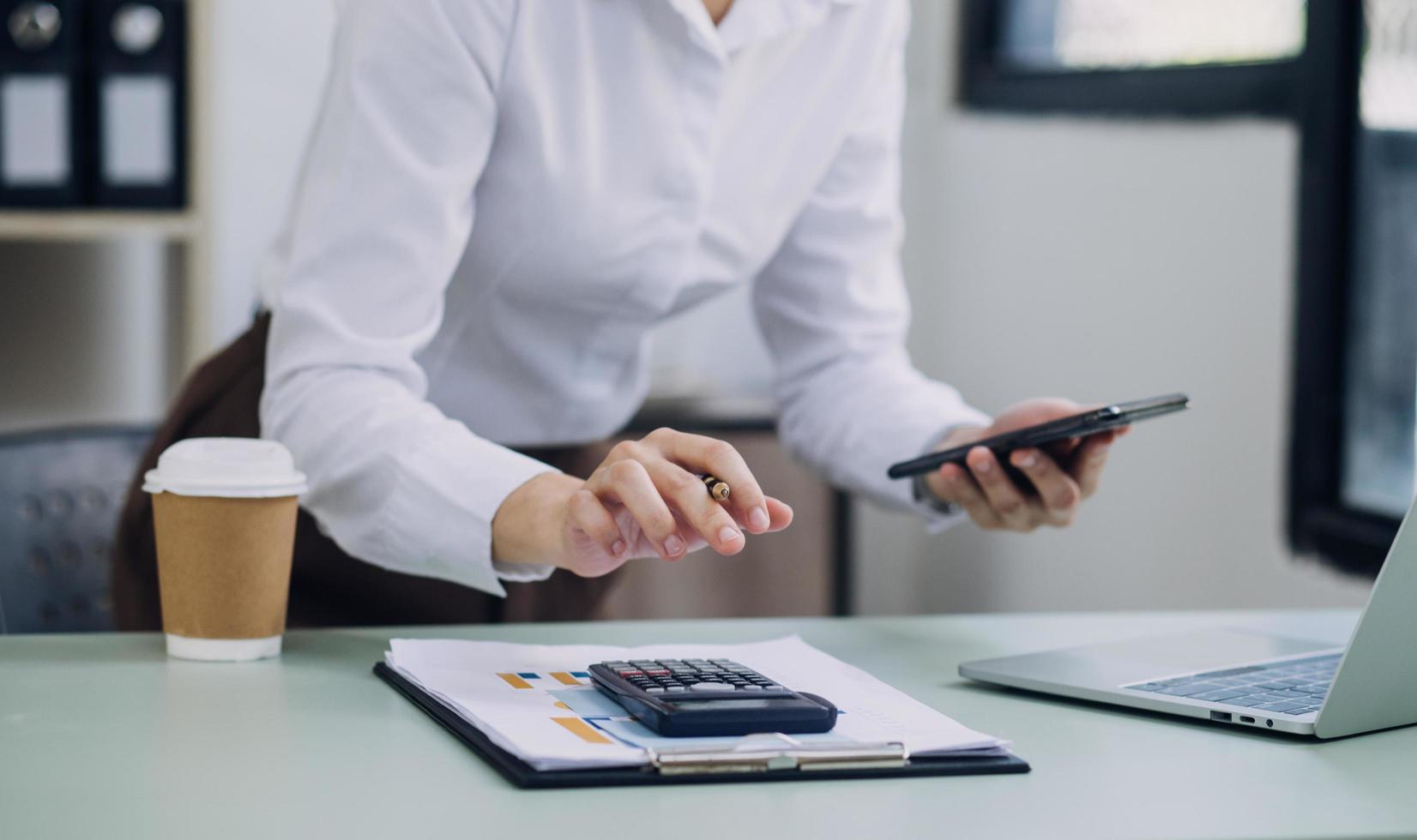 Young business woman sitting in office at table and using smartphone. On desk is laptop and tablet computer, on screen charts and graphs. Woman analyzing data. Student learning online. photo