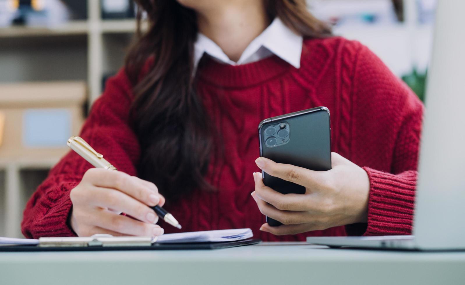 Young business woman sitting in office at table and using smartphone. On desk is laptop and tablet computer, on screen charts and graphs. Woman analyzing data. Student learning online. photo