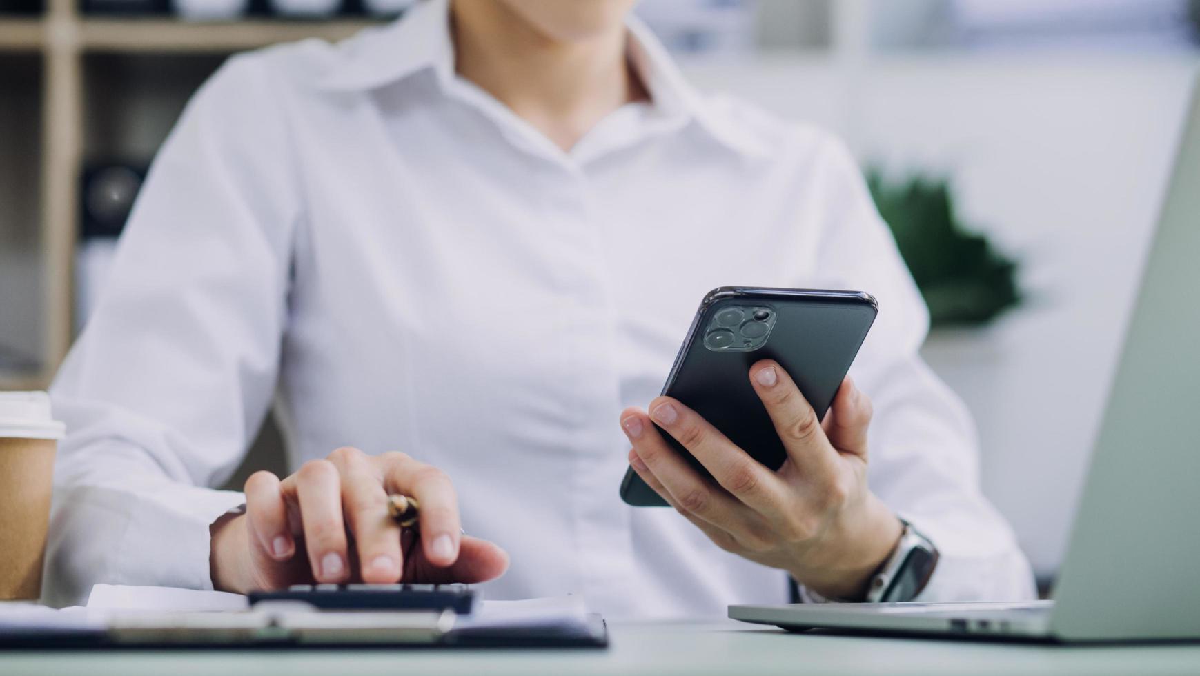 Young business woman sitting in office at table and using smartphone. On desk is laptop and tablet computer, on screen charts and graphs. Woman analyzing data. Student learning online. photo