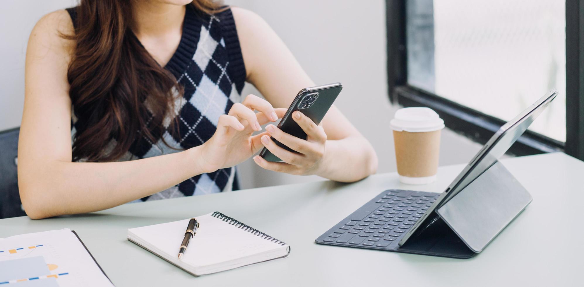 Young business woman sitting in office at table and using smartphone. On desk is laptop and tablet computer, on screen charts and graphs. Woman analyzing data. Student learning online. photo