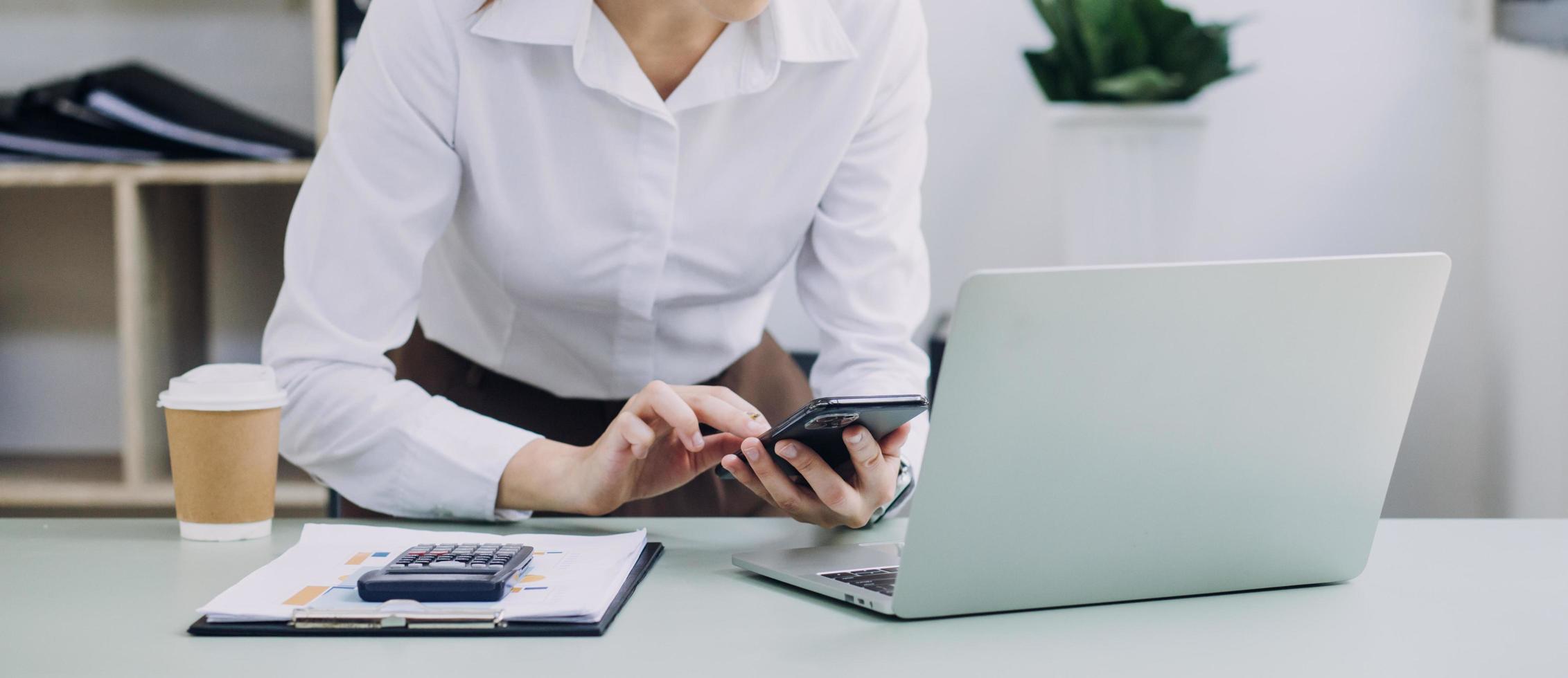 Young business woman sitting in office at table and using smartphone. On desk is laptop and tablet computer, on screen charts and graphs. Woman analyzing data. Student learning online. photo