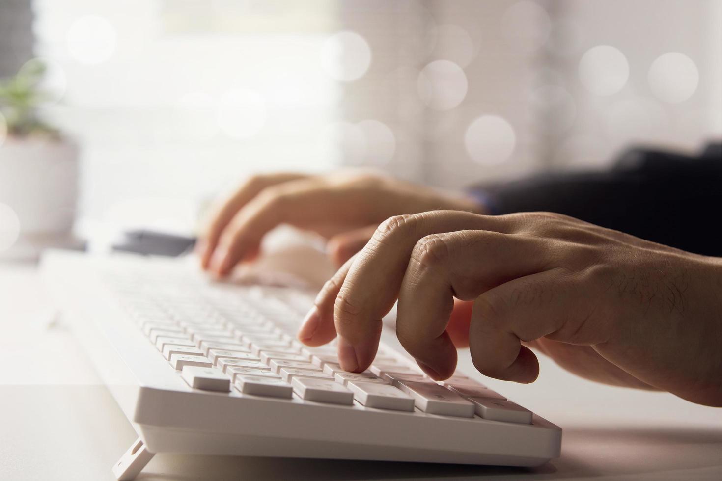 side view man hands typing computer keyboard White laptop on desk with blurry light illuminating window in office. Workspace. Freestanding office corner. photo