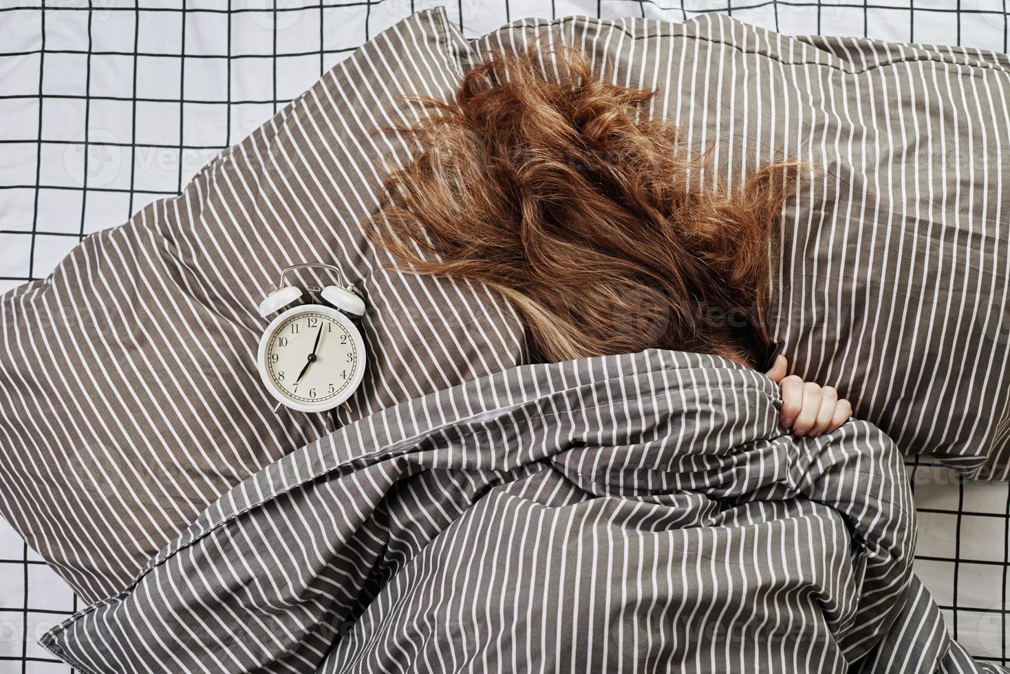 mujer dormida cubierta bajo una manta en la cama y reloj despertador vintage en la almohada foto