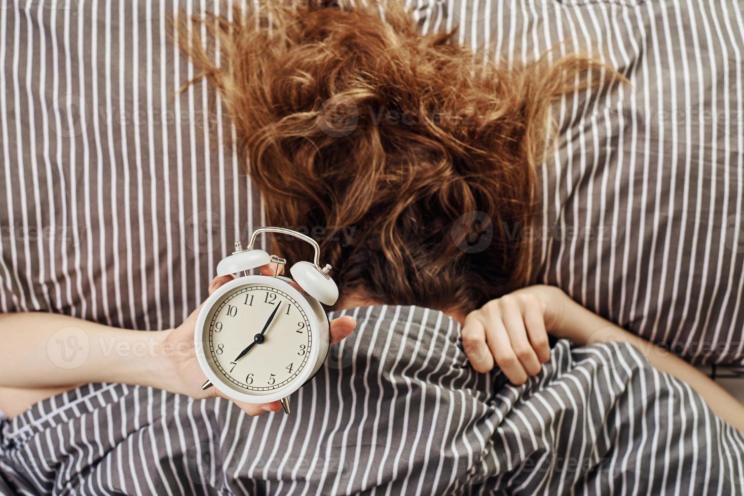 mujer dormida cubierta bajo una manta en la cama sostenga el despertador en la mano foto