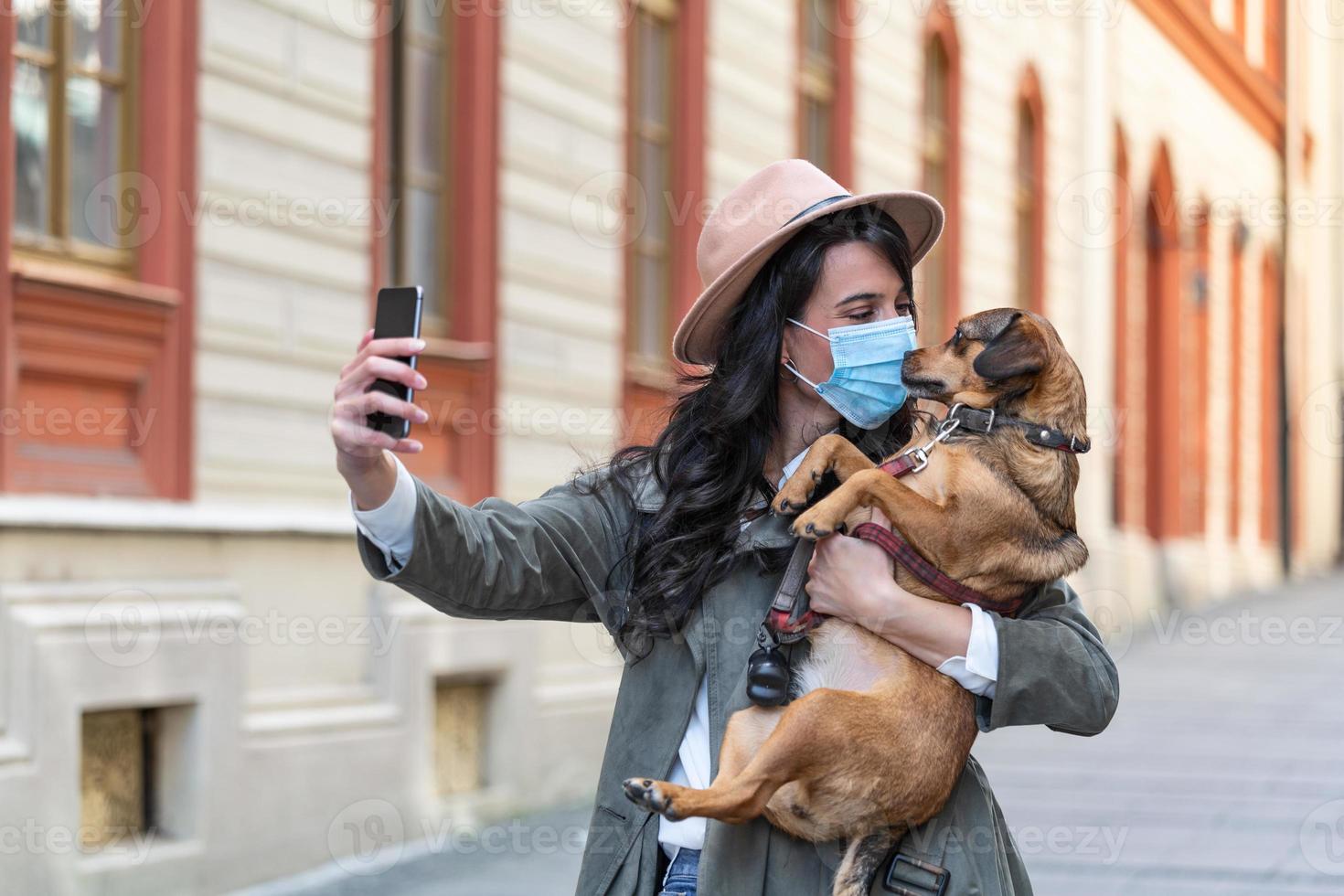 Woman during pandemic isolation walking with her dog and using mobile phone to make a selfie. Stylish girl with funny dog resting, hugging and having fun, cute moments. photo