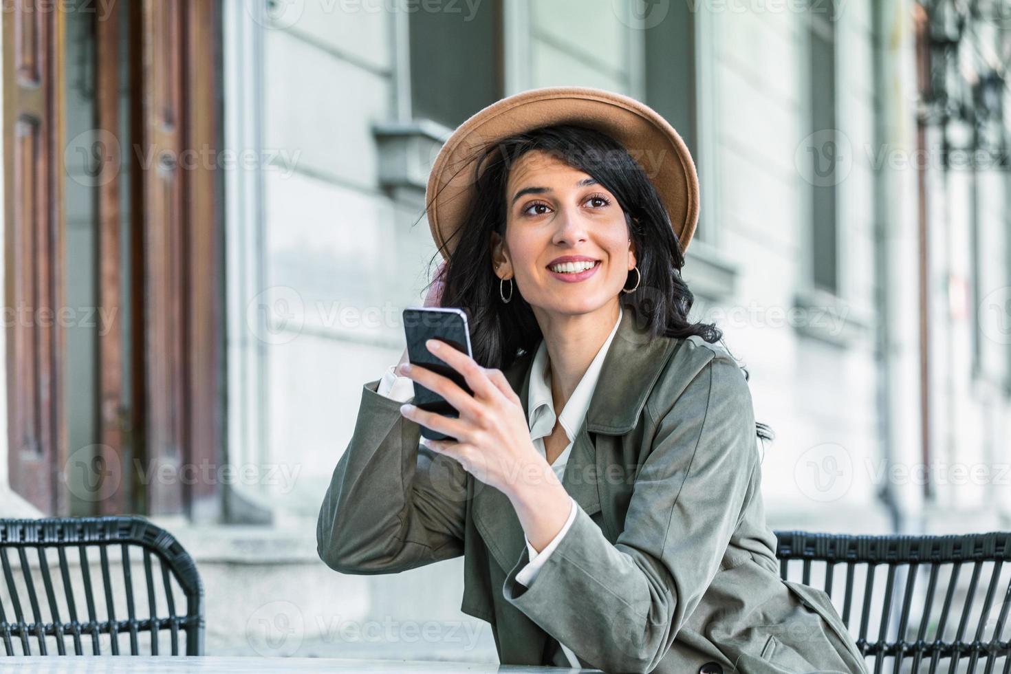 mujer hermosa joven mirando el teléfono inteligente mientras está sentado en la cafetería. estudiante universitario feliz usando teléfono móvil. mujer de negocios tomando café, sonriendo y usando un teléfono inteligente en el interior. foto